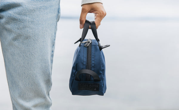 A person holding a Navy Parapack Grab Bag by it's grab handle showing the TOM BIHN Design Lab label.